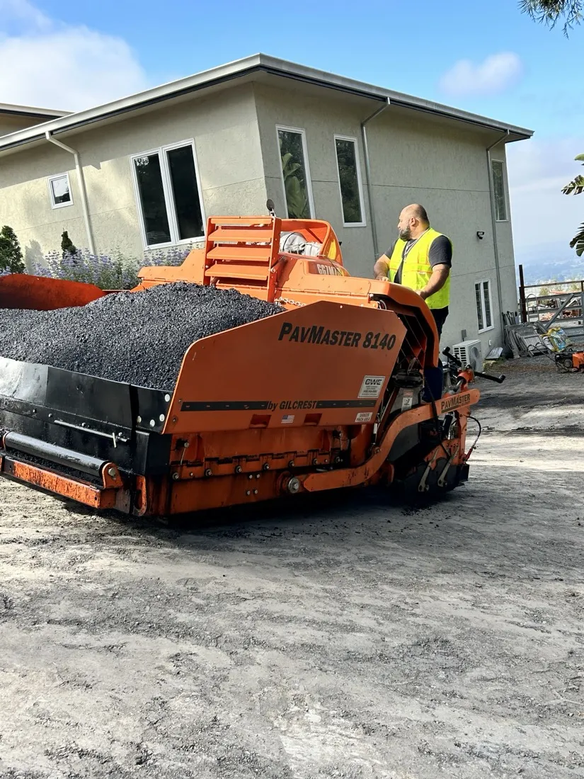 Asphalt paver with a loaded hopper on a graded base beside a hillside home