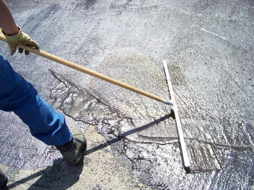 Worker spreading liquid sealer across asphalt with a long-handled squeegee